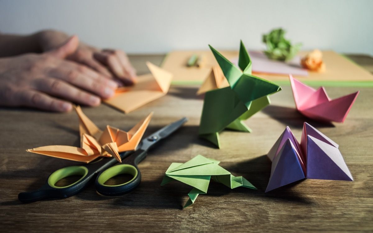 Nekoliko figurica origami na drvenom stolu, u pozadini rukama presavijajući papir u boji.