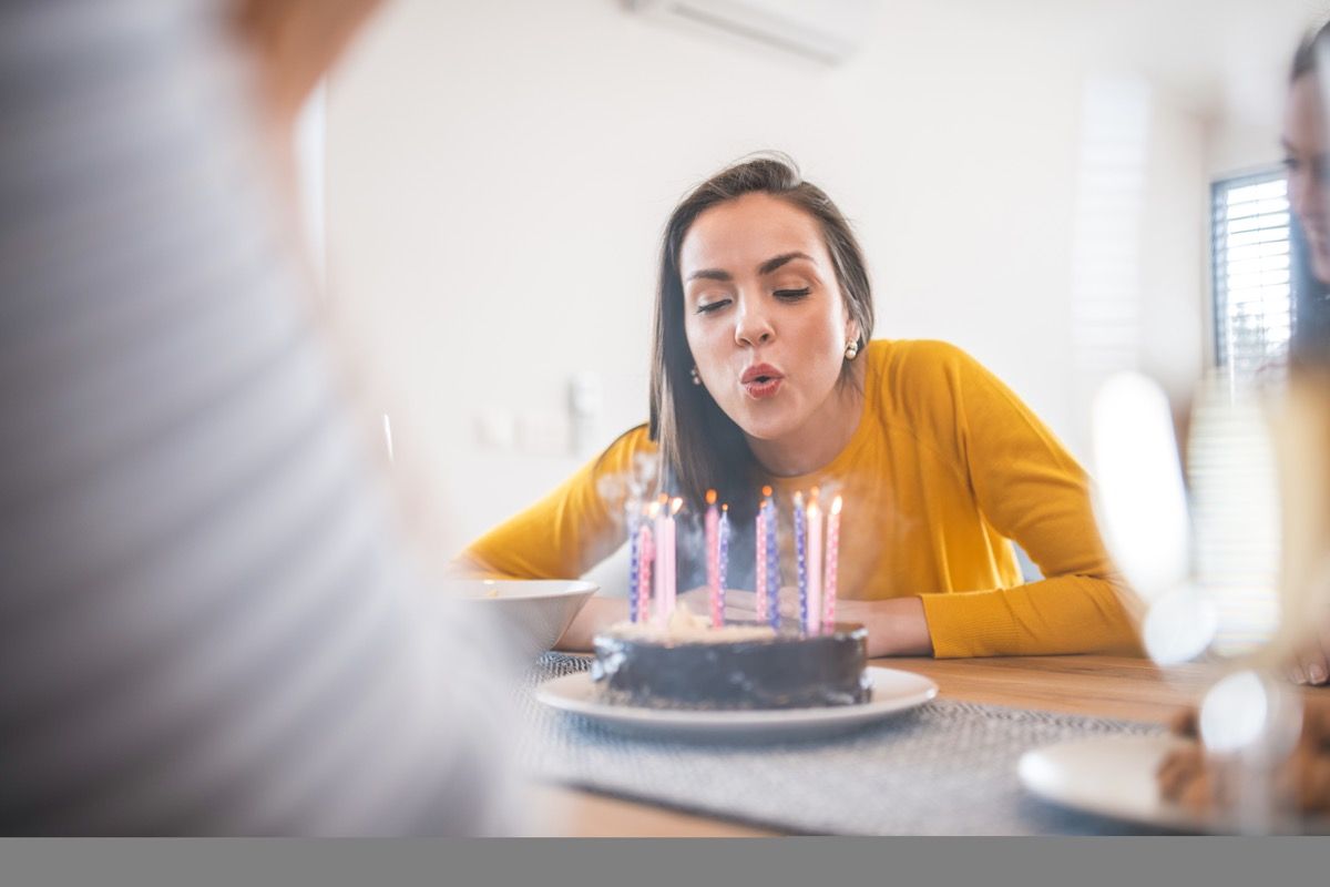 Kaunis nuori nainen puhaltaa kynttilöitä kakkuun ruokapöydässä. Naiset juhlivat ystävänsä syntymäpäiväjuhlia. He ovat kotona.