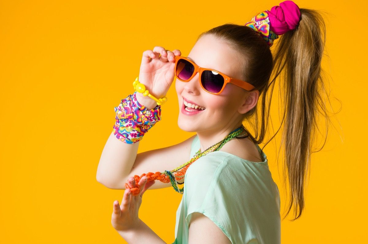 Besišypsanti mergina su spalvingais drabužiais, akiniais nuo saulės, geltonu fonu.