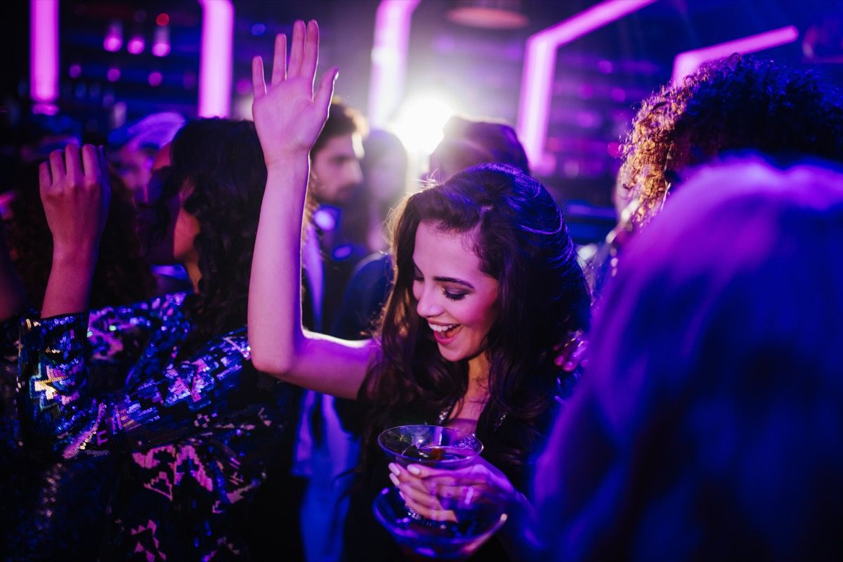 Wanita dewasa muda yang bahagia menari bersama teman-temannya di klub malam sambil memegang koktail di tangan