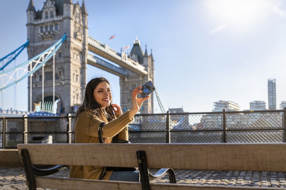 सुंदर महिला लंदन यात्री एक धूप के दिन टॉवर ब्रिज के सामने अपने फोन के साथ एक सेल्फी तस्वीर लेती है