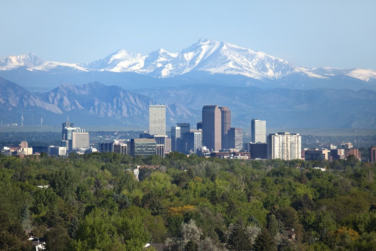 Lumipeitteinen Longs Peak, osa Kalliovuoria, on korkealla taustalla vihreillä puilla ja Denverin keskustan pilvenpiirtäjillä sekä hotelleilla, toimistorakennuksilla ja kerrostaloilla, jotka täyttävät taivaanrantaa.