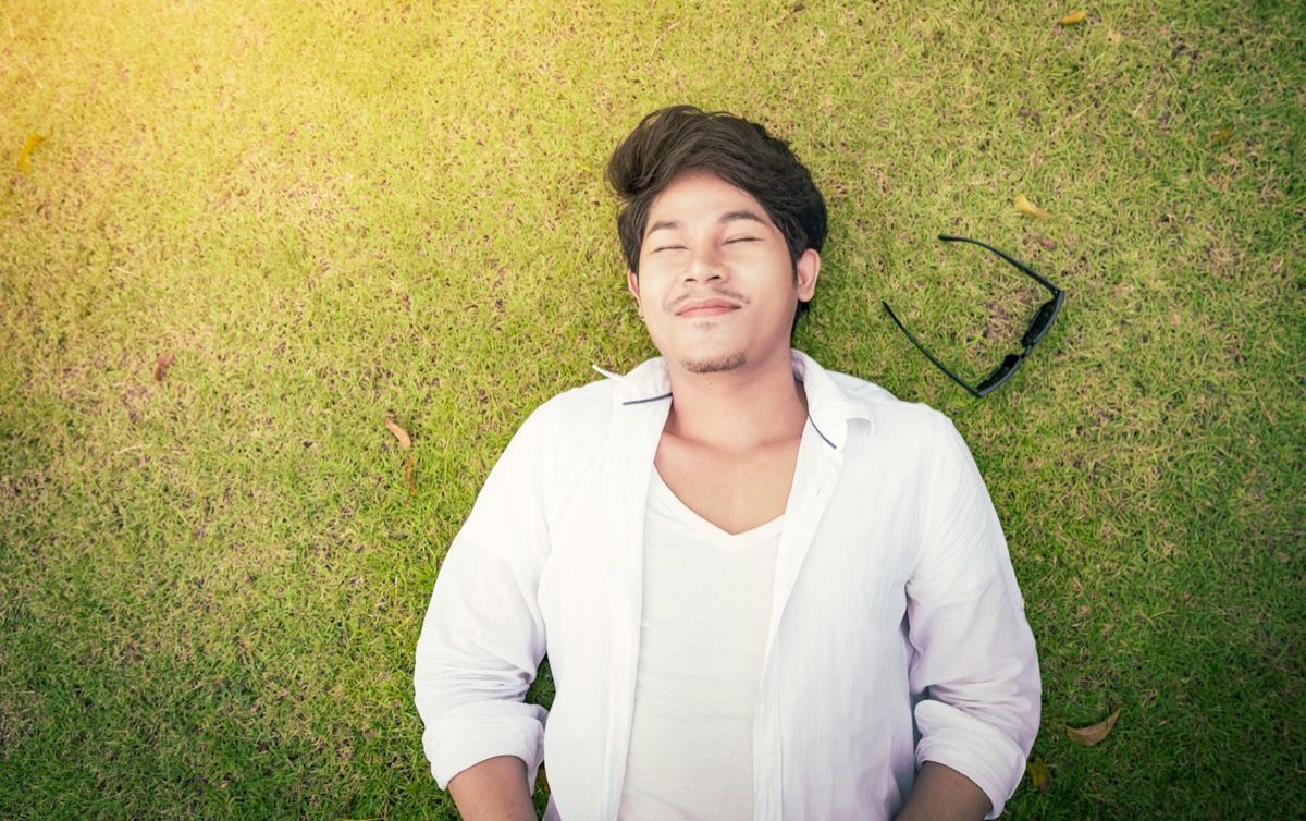 млад азиатски мъж, лежащ на тревата със затворени очи и слънчеви очила до себе си