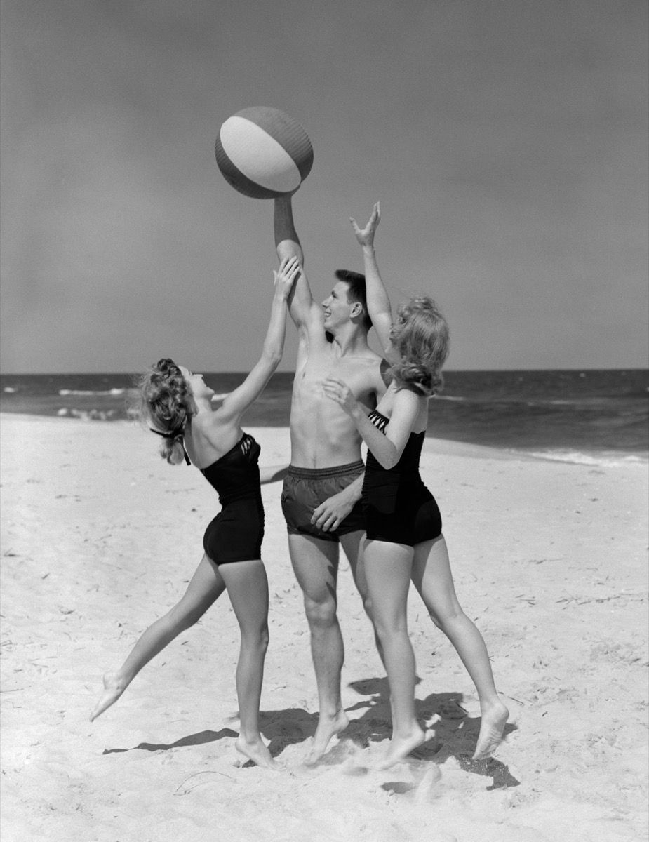 Две тинејџерке из 1950-их посежу за лоптом на плажи у мушким тинејџерима на рукама на плажи, хладне баке и деке