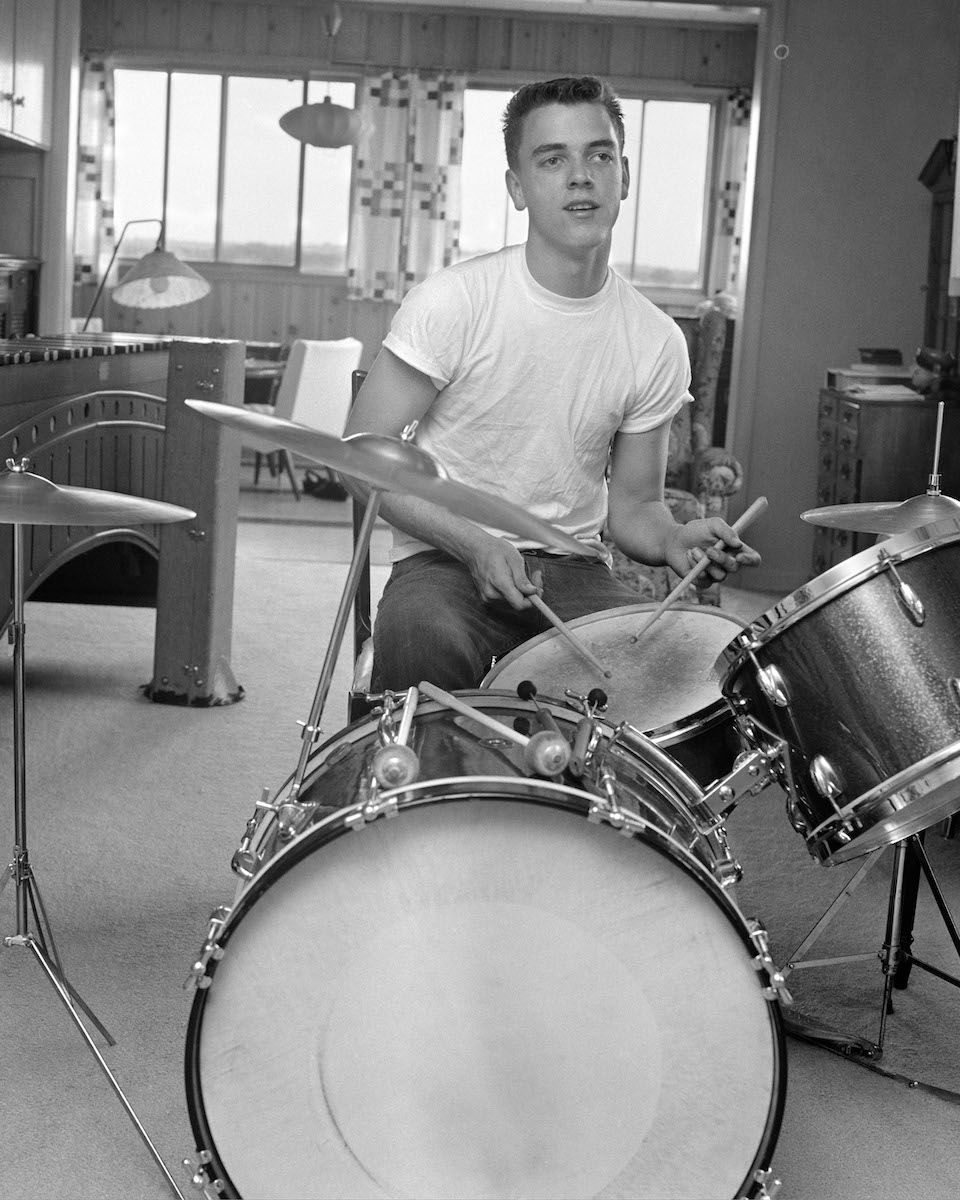 1960ndate aastate teismeline poiss, kes oli lõigatud BUZZiga T-särkides, istudes väikeste trummikomplektide ees, millel on üle lõhe