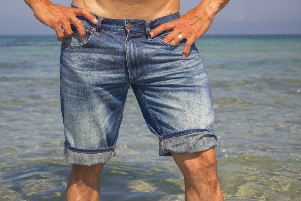 Homem vestindo shorts jeans em pé na água do mar, closeup de pernas