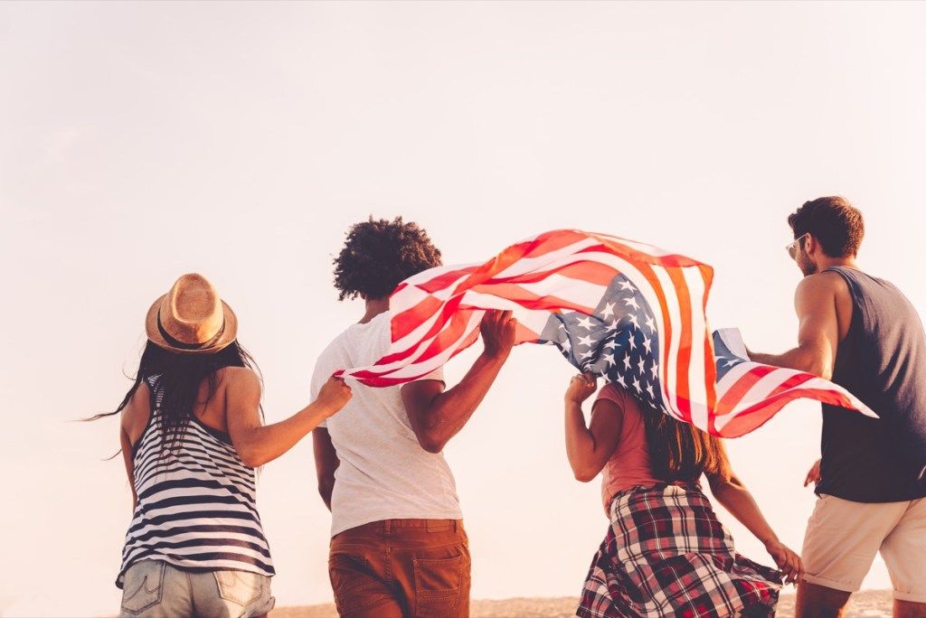 Приятели с американско знаме. Изглед отзад на четирима млади хора, носещи американски флаг, докато тичат на открито - Изображение