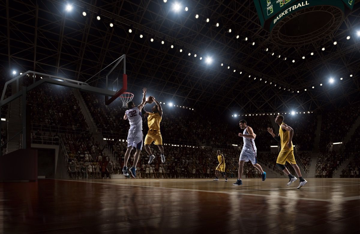 Pogled na profesionalno košarkarsko tekmo z nizkim kotom. Igralec v zraku drži žogo, ki bo kmalu dosegel slam dunk, toda igralec iz nasprotne ekipe ga je pripravljen blokirati. Igra se v zaprtem osvetljenem košarkarskem prostoru. Vsi igralci nosijo generično košarkarsko uniformo brez blagovne znamke.