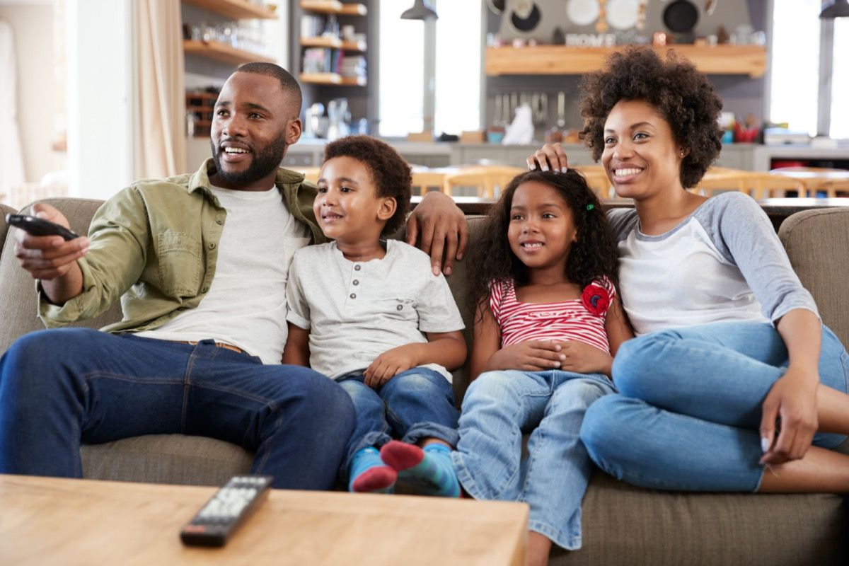 jaunas juodaodis vyras, moteris ir du vaikai, sėdintys ant sofos ir žiūrėdami televizorių