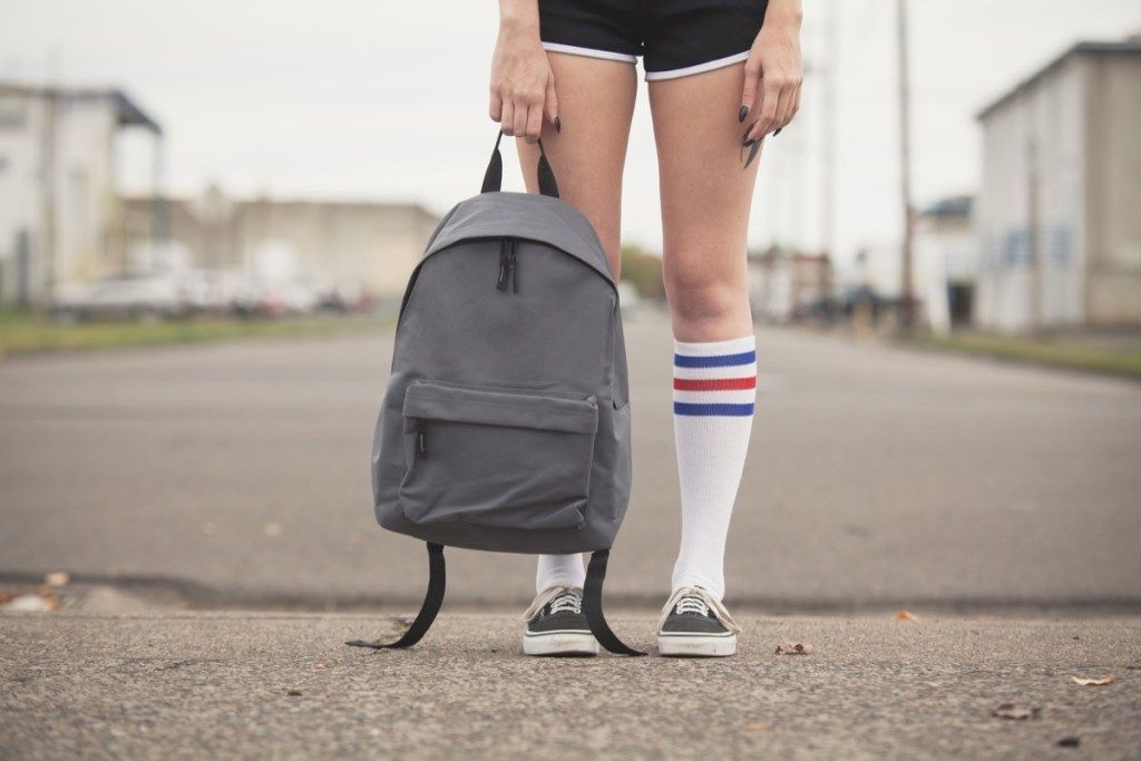 Dekle drži generični nahrbtnik v črnih kratkih hlačah in nogavici s teniškimi čevlji na prosti ulici. - Slika