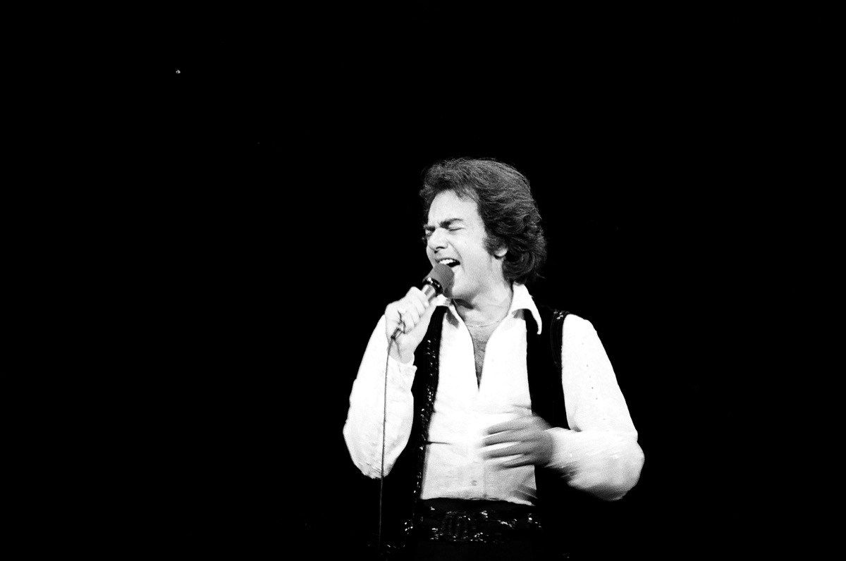 Neil Diamond nastopa v beli srajci s širokim ovratnikom in črnem jopiču