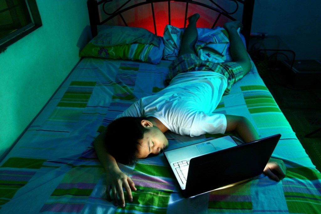 тийнейджър, заспал с лице нагоре пред все още включен лаптоп и изпълняващ домашни