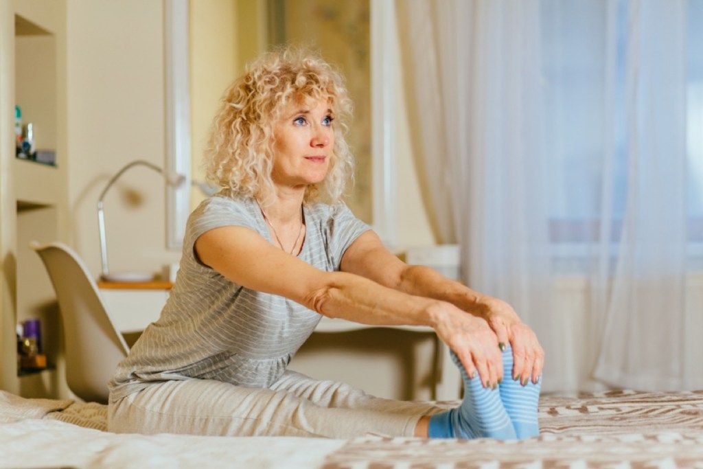 keski-ikäinen valkoinen nainen venyttää sängyssä pitämällä kiinni jaloistaan