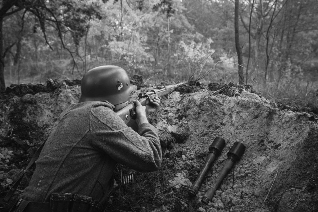 Nežinomas atkūrėjas, apsirengęs vokiečių Wehrmacht pėstininkų kariu Antrojo pasaulinio karo metais, paslėptas sėdėdamas su šautuvo ginklu pasaloje tranšėjoje rudens miške. Nuotrauka juodos ir baltos spalvos.