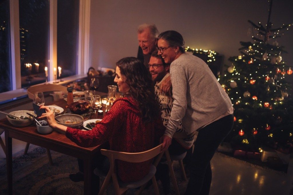 Štiri osebe za mizo pred božičnim drevescem in ena ženska, ki drži telefon