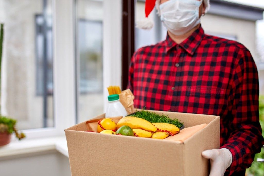 Un hombre que sostiene una caja con comida mientras usa una máscara y un gorro de Papá Noel