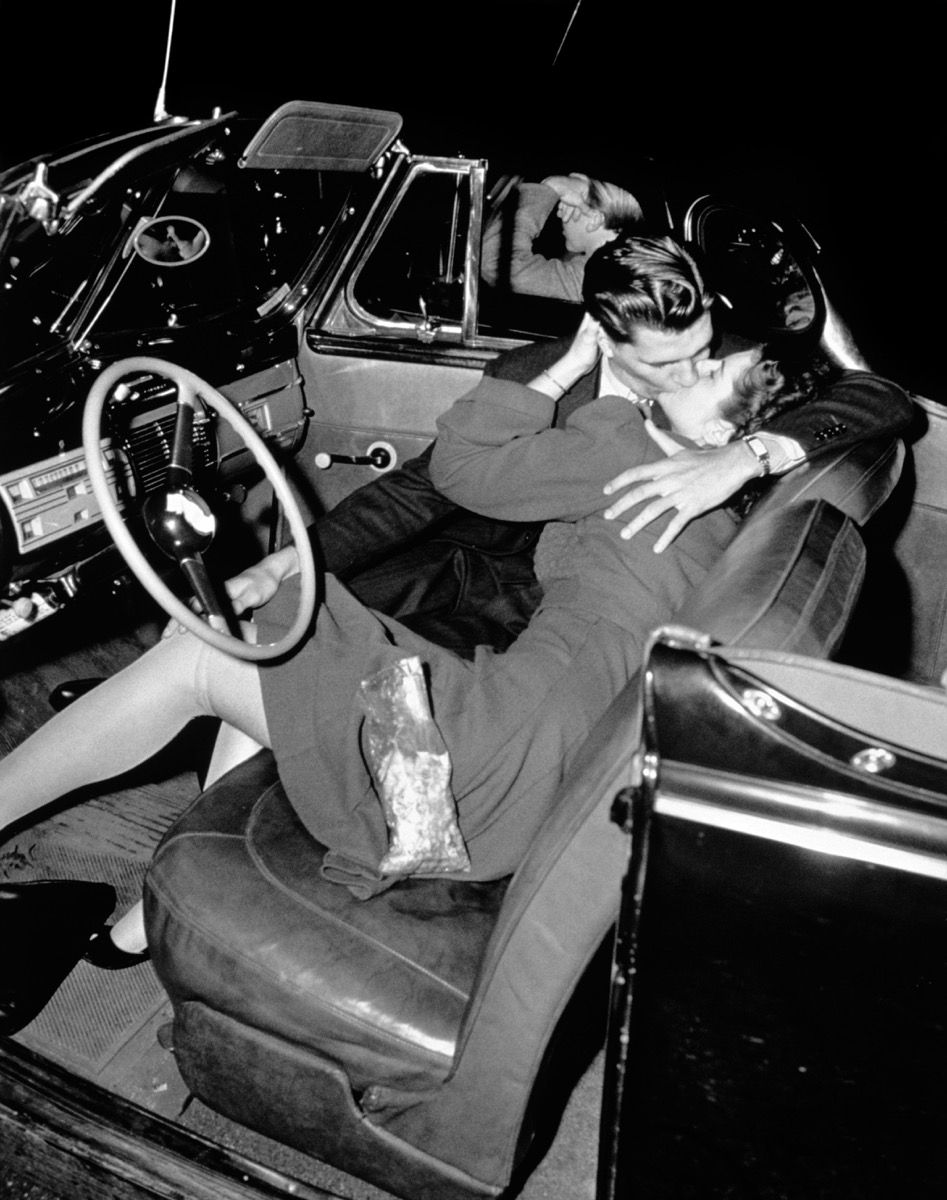 Pāris, kurš skūpstās savā automašīnā pie iebraukšanas 1950. gados {Iepazīšanās pirms 50 gadiem}