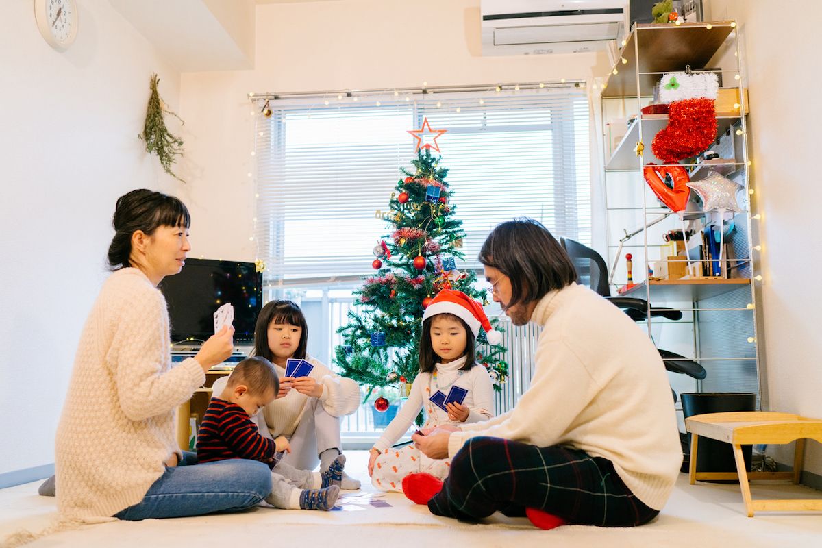 Una familia está sentada en el suelo y jugando a las cartas en la sala de estar decorada con adornos navideños y un árbol de Navidad durante la Navidad.