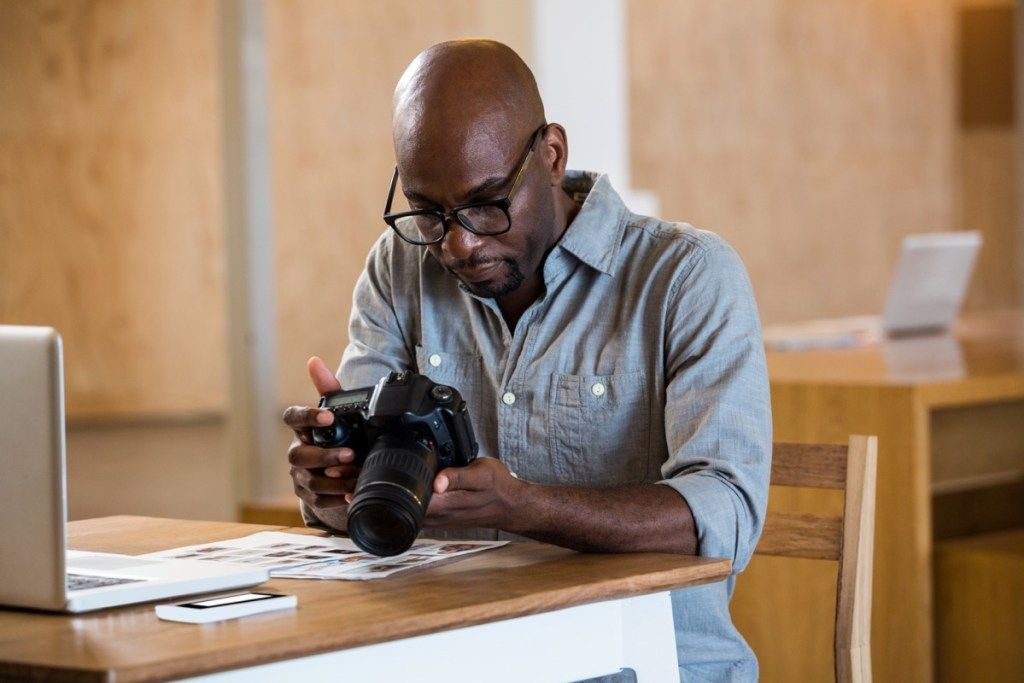 muž v džínsovej košeli s čiernymi okuliarmi drží fotoaparát pred počítačom