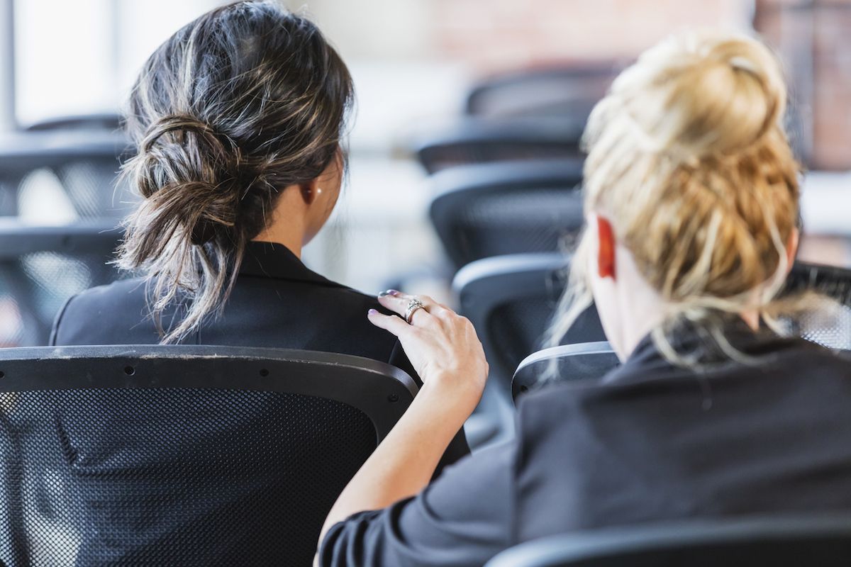 As costas das cabeças de duas empresárias sentadas em cadeiras, participando de uma conferência ou seminário de negócios. Uma mulher está batendo no ombro da outra tentando chamar sua atenção.