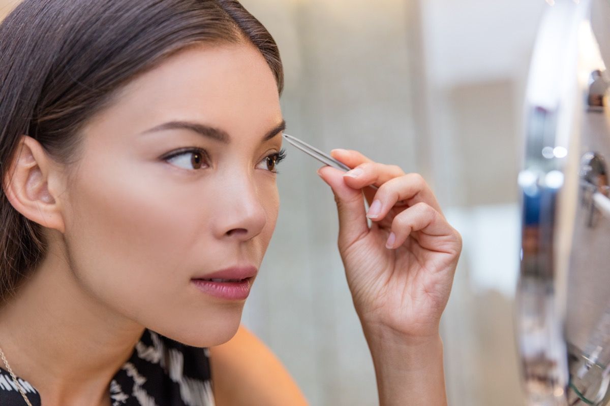 Aziatische vrouw wenkbrauwen plukken met een pincet met behulp van een wenkbrauwpincet thuis in de badkamer make-up spiegel. Close-up van een meisje