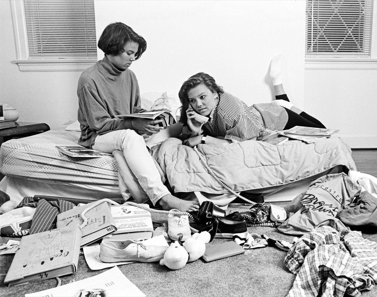 Divas pusaudžu afrikāņu amerikāņu meitenes pārblīvētā guļamistabā, kas runā pa tālruni un vienu lasāmo žurnālu