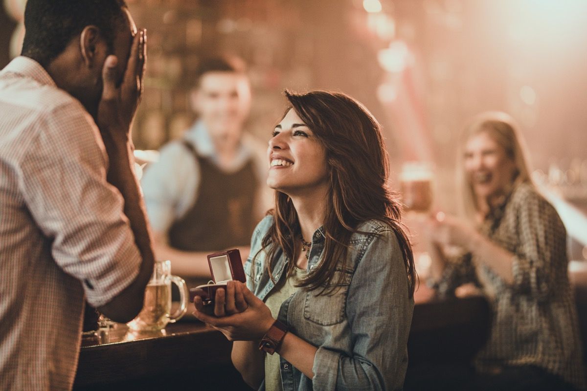 Mujer joven feliz proponiendo a su novio negro durante la noche en un pub. Hay gente al fondo.