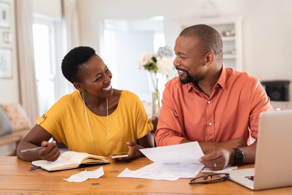 Veselý dospelý pár sedí a spravuje výdavky doma. Šťastný africký muž a žena spolu platia účty a riadia rozpočet. Čierny usmievavý pár kontrolujúci účtovníctvo a účty pri pohľade na seba.