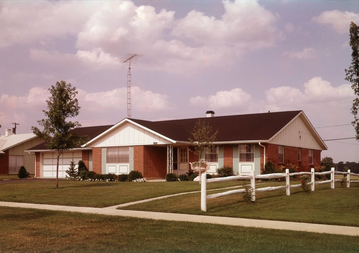 บ้านไร่ชานเมืองขนาดใหญ่ในปี 1960 ตั้งอยู่บนพื้นหญ้าสีเขียวพร้อมรั้วล้อมรั้วสีขาว