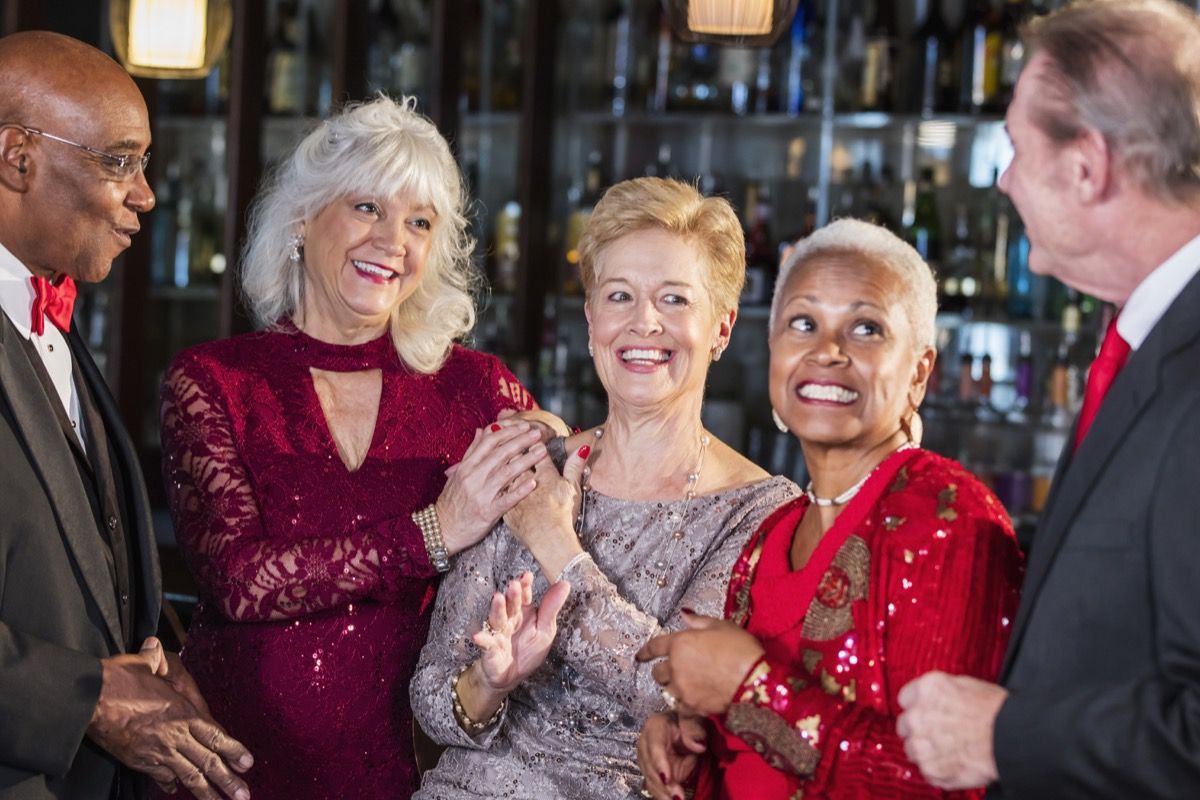 Eine multiethnische Gruppe von fünf Senioren genießt einen Abend, hängt zusammen an der Theke eines Restaurants, redet und lacht. Sie sind gut gekleidet und tragen Anzüge und Kleider. Die Frau in der Mitte ist in den 70ern und ihre Freunde sind in den 60ern.