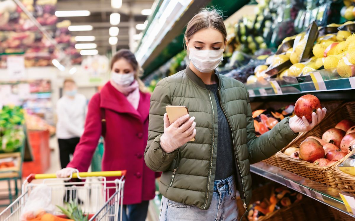 Млада жена с маска за лице за еднократна употреба проверява списък за пазаруване на смартфон, докато има друга жена с фона на количката за пазаруване