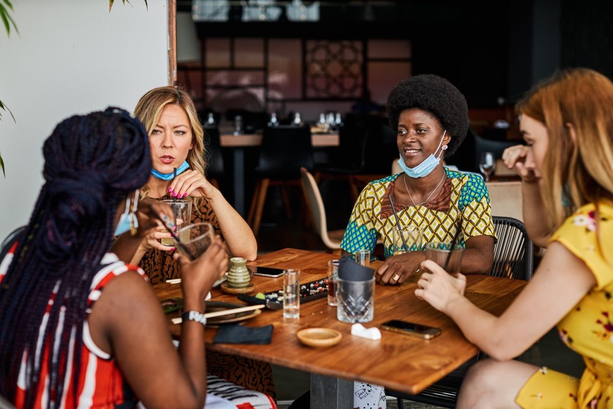 Neljä nuorta naista istuu ravintolassa, yllään kasvonaamio leuassa.