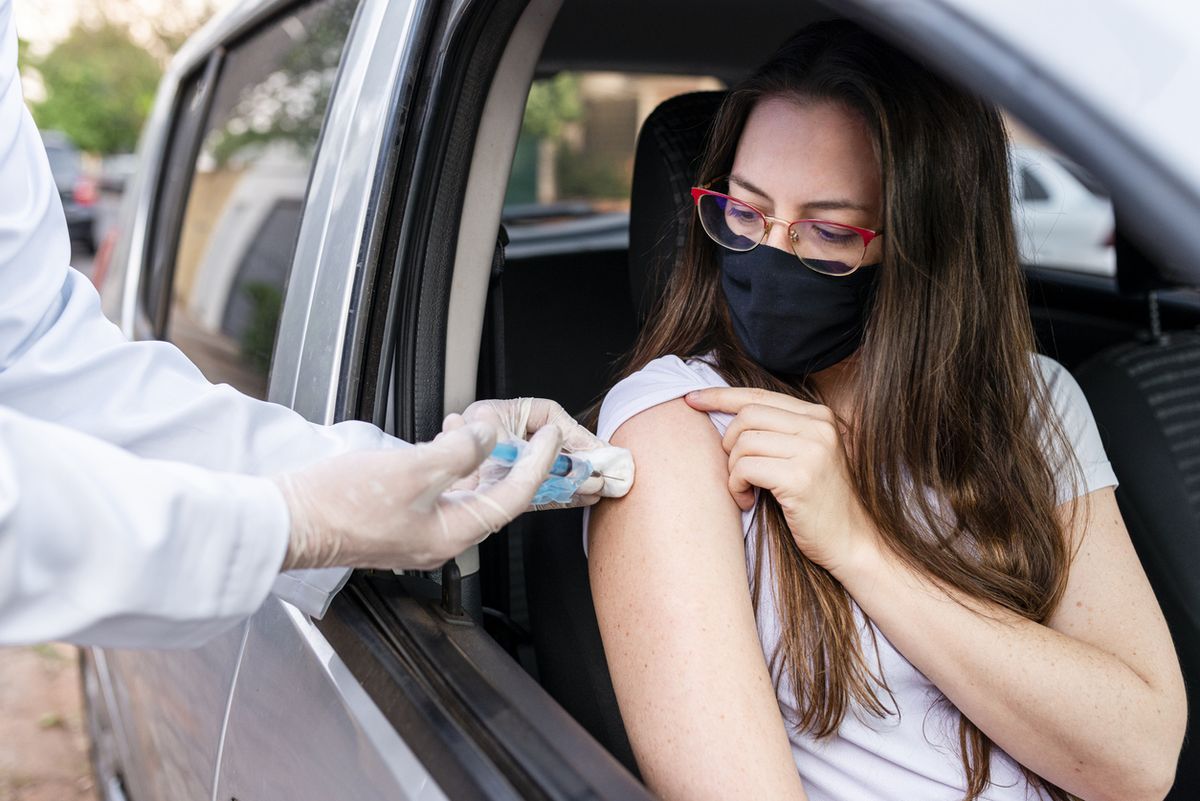 Mladá žena sediaca v aute a oblečená v maske dostane vakcínu COVID-19 od zdravotníckeho pracovníka v rukaviciach.
