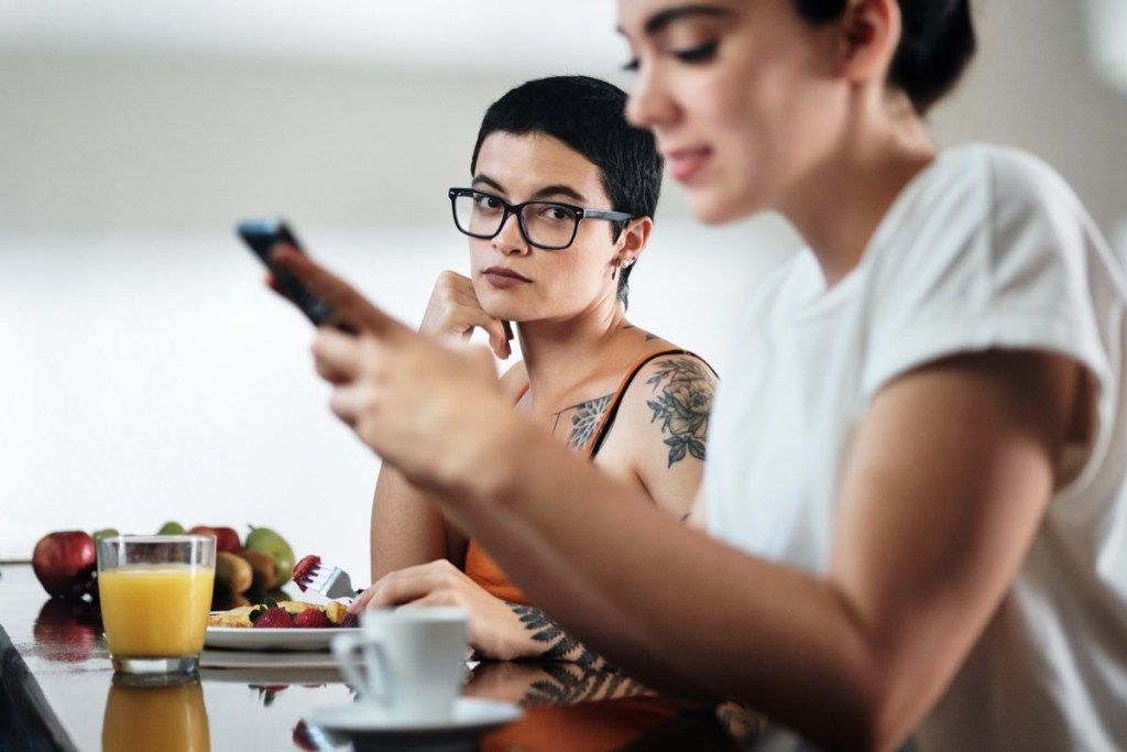 kaksi lesboa naista kotona syömässä aamiaista, kumppani juttelemassa matkapuhelimella. Tyttöystävä jättää nuoren naisen huomiotta ja tuntee mustasukkaisuuden