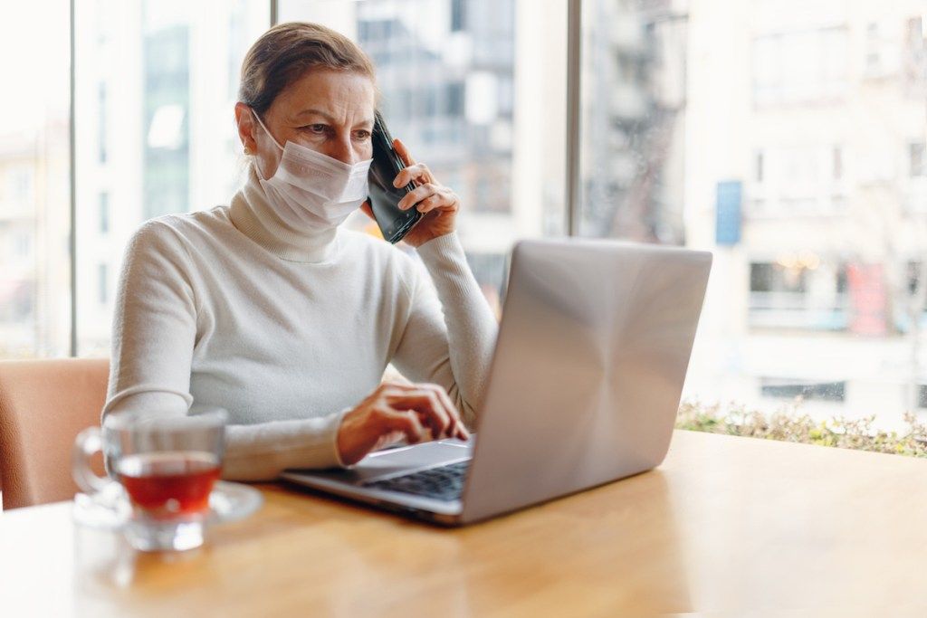 femeie senior cu mască de față, stând la computer în timp ce stă la telefon
