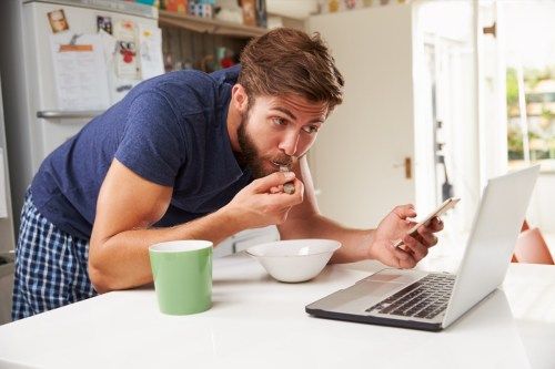 איש ריבוי משימות במטבח אוכל, עומד, צופה במחשב נייד, מחזיק טלפון