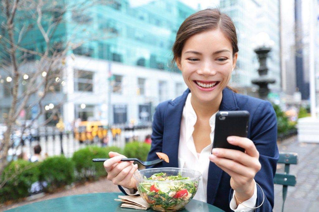 אישה מחייכת בזמן שהיא אוכלת סלט לארוחת הצהריים לבדה ומביטה בטלפון שלה