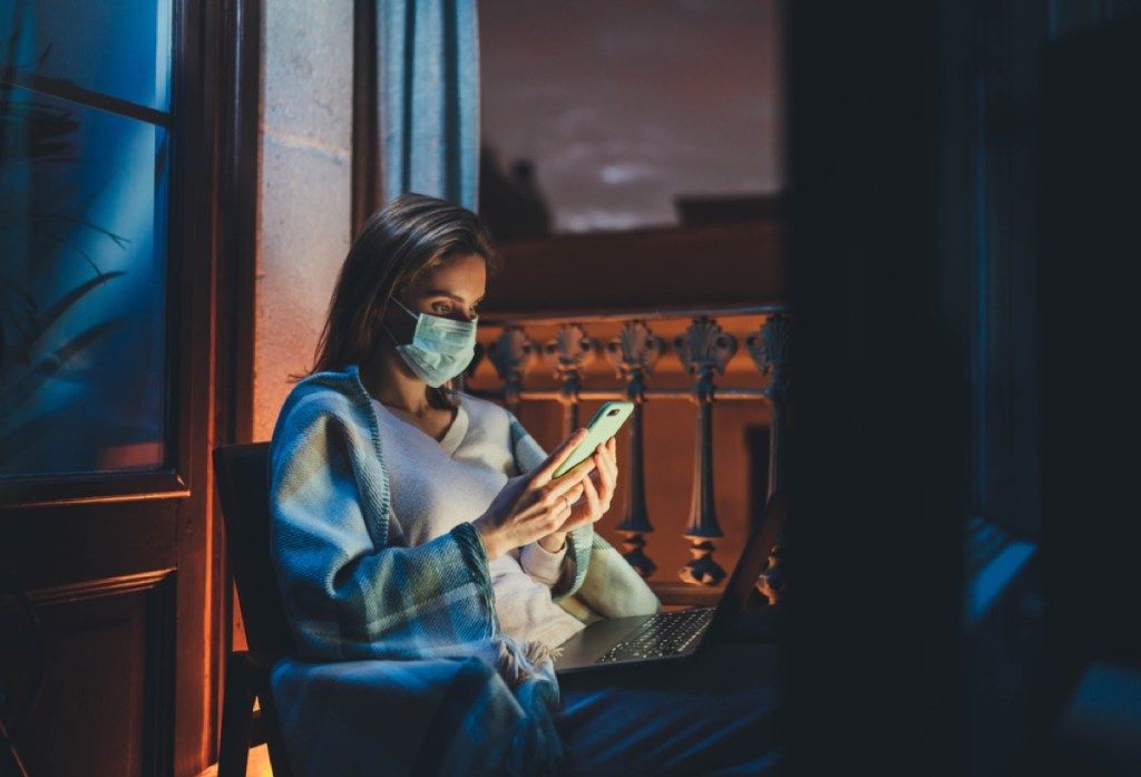 жена с маска за лице, която седи на прозореца и гледа телефона и компютъра си