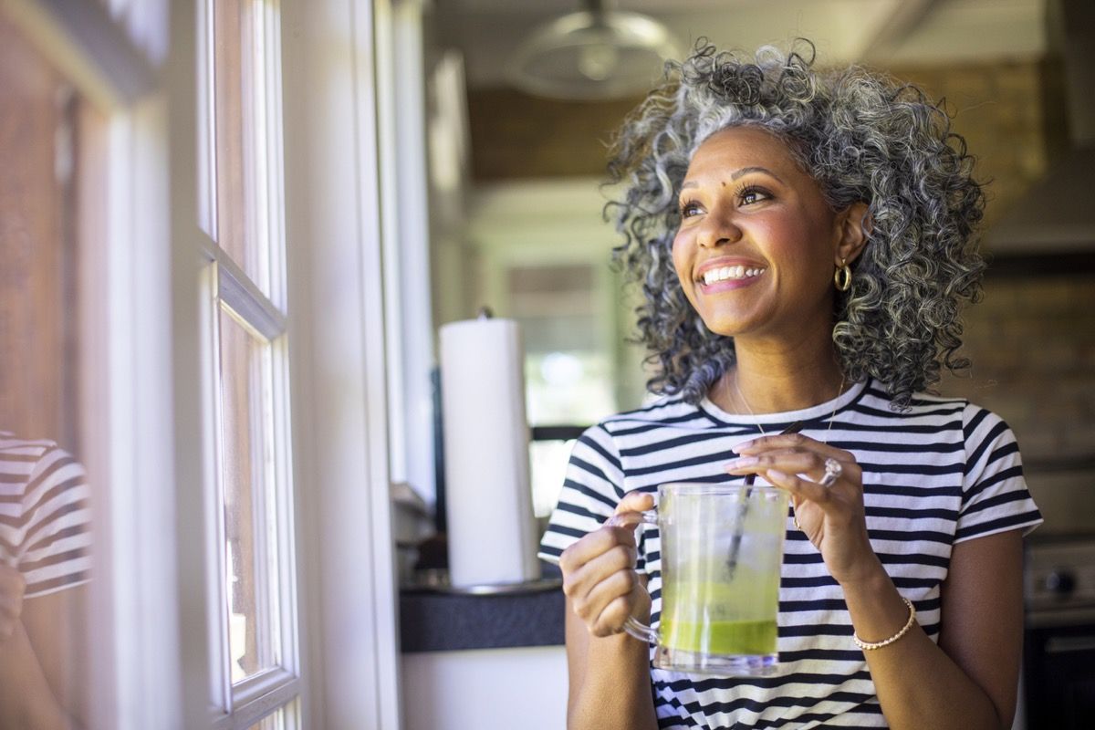 Mujer negra de mediana edad con cabello blanco y rizado bebe un batido verde en la mañana para el desayuno