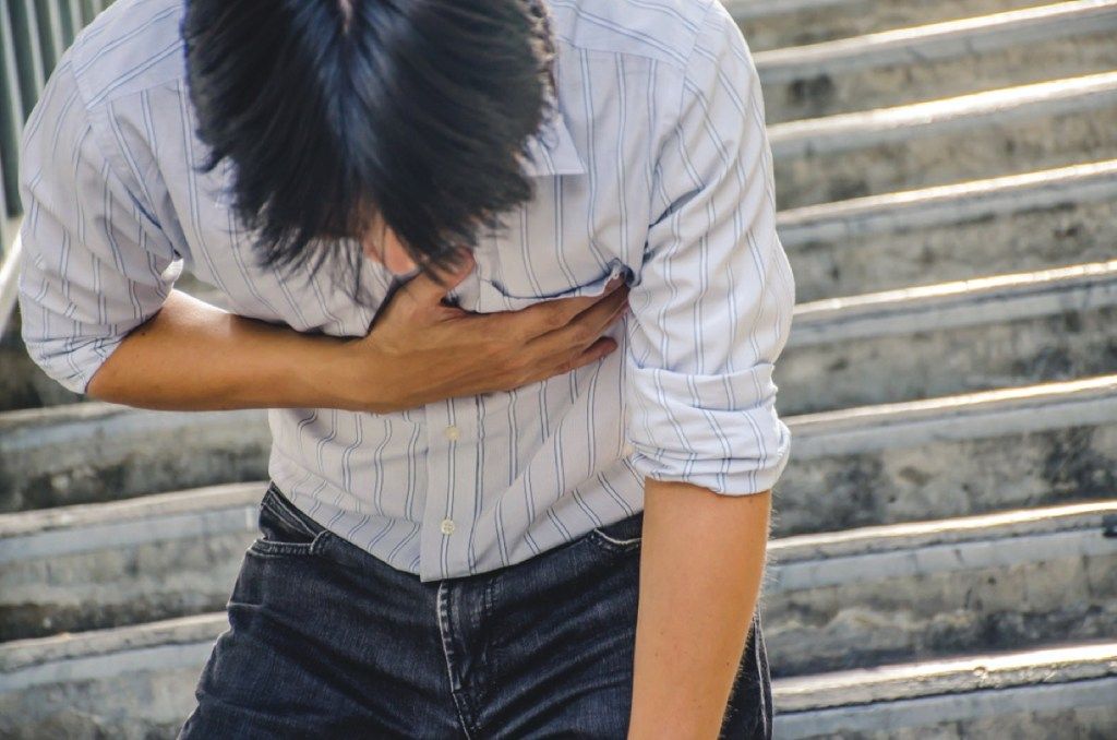 युवा एशियाई आदमी सीढ़ियों पर छाती को पकड़ता है, संकेत देता है कि आपकी सर्दी गंभीर है