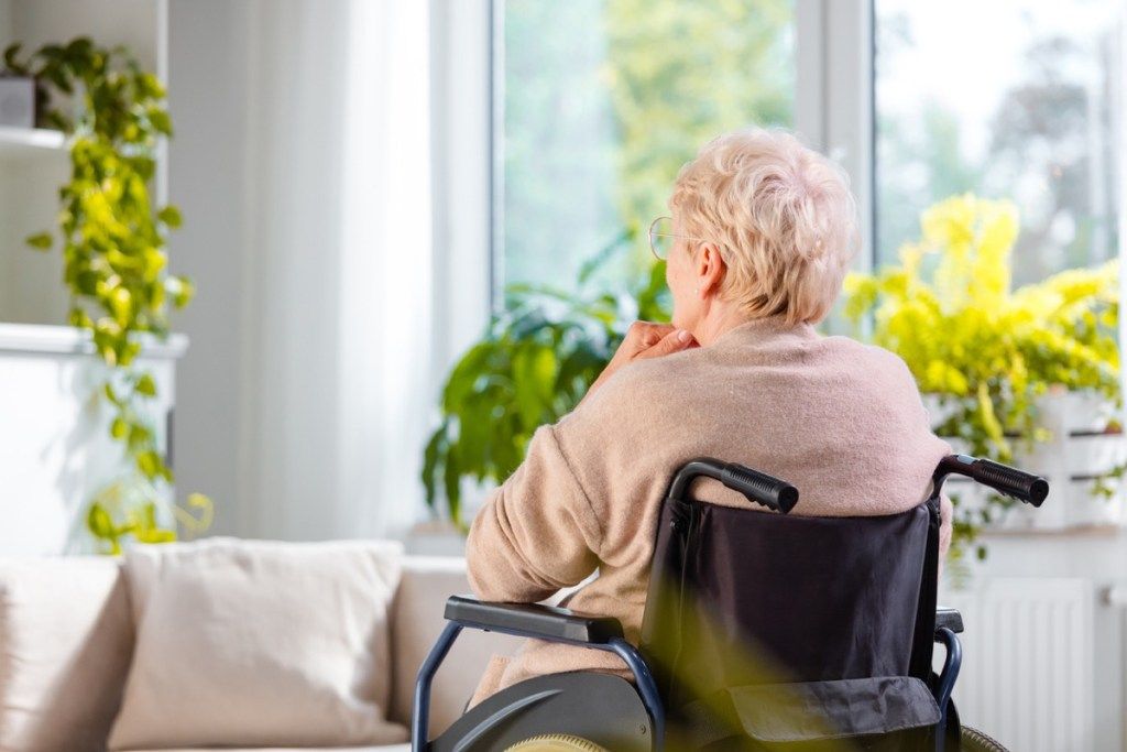 Galinio vaizdo kadras, kuriame matoma vyresnio amžiaus moteris, sėdinti invalido vežimėlyje ir mąstanti