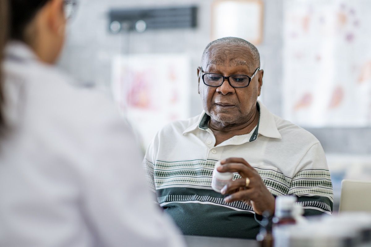 एक बुज़ुर्ग अश्वेत व्यक्ति डॉक्टर के एक डॉक्टर से मेज के पार बैठकर अपने हाथ में एक गोली की बोतल देखता है