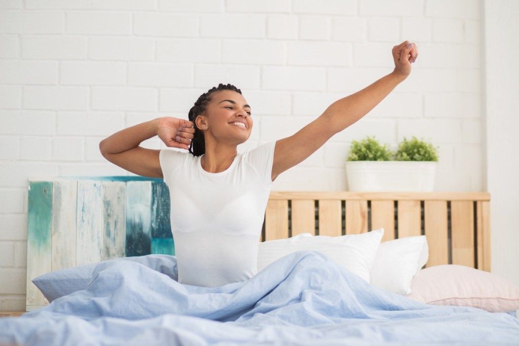 젊은 흑인 여성이 아침에 흰색 티셔츠를 입고 침대에 앉아 뻗어, 놀라운 느낌을주는 방법