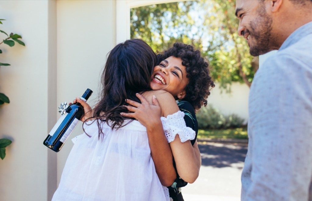Schwarze Frau mit natürlichem Haar umarmt Freundin an der Tür mit Einweihungswein in der Hand, Möglichkeiten, sich erstaunlich zu fühlen