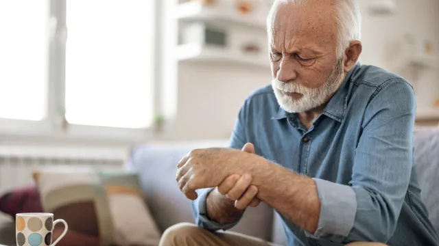   Vecāks vīrietis, kas sēž uz dīvāna un berzē roku artrīta dēļ.