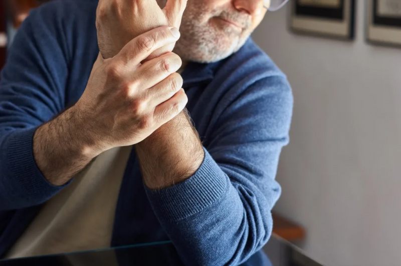   vīrietis sēž mājās, turot kreisās rokas plaukstas locītavu osteoartrīta, artrīta, tendinīta, reimatisma izraisītu sāpju dēļ.