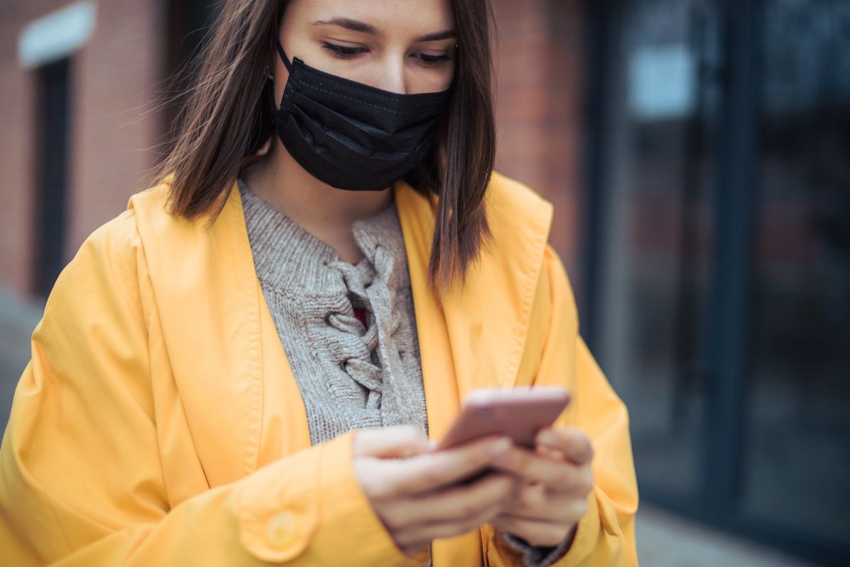 Jauna sieviete, kurai ir dzeltena jaka un sejas maska, pārbauda savu viedtālruni.