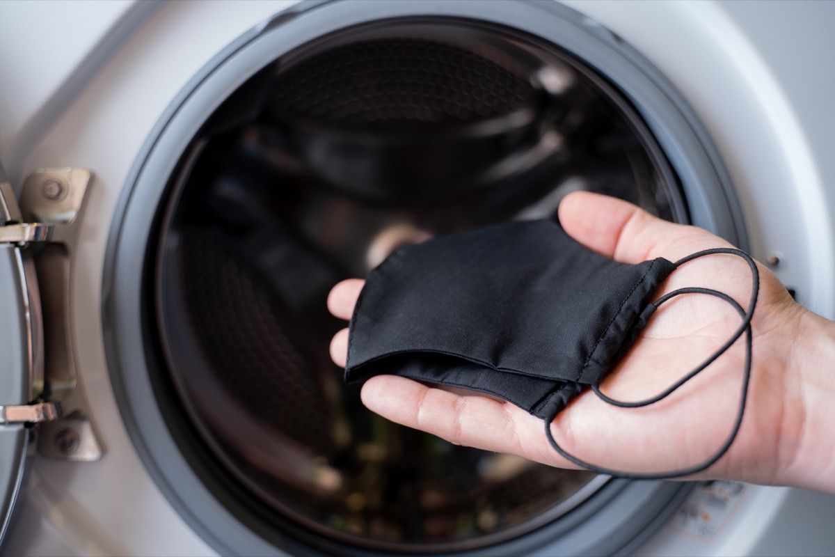 एक व्यक्ति का हाथ जो वॉशिंग मशीन में एक काला मुखौटा फेंक रहा है