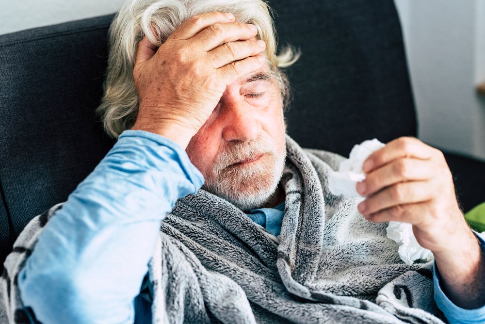 Пожилой мужчина сидит на кушетке, завернувшись в одеяло, прикасаясь ко лбу и чувствуя жар, страдая от симптомов COVID.