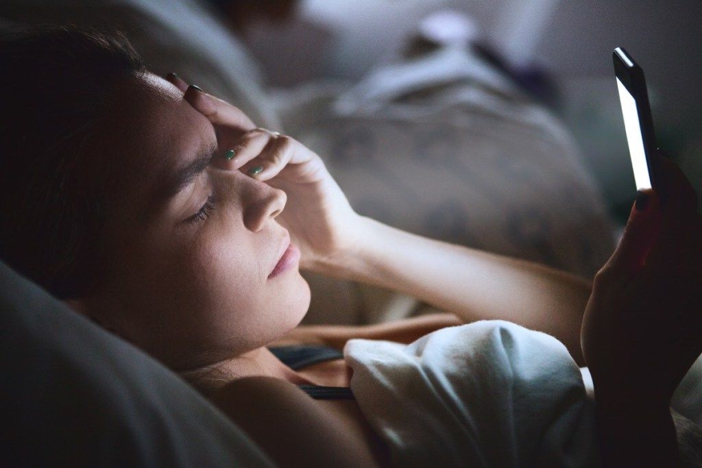 Nainen makaa sängyssä puhelinta käyttäen päänsärkyä